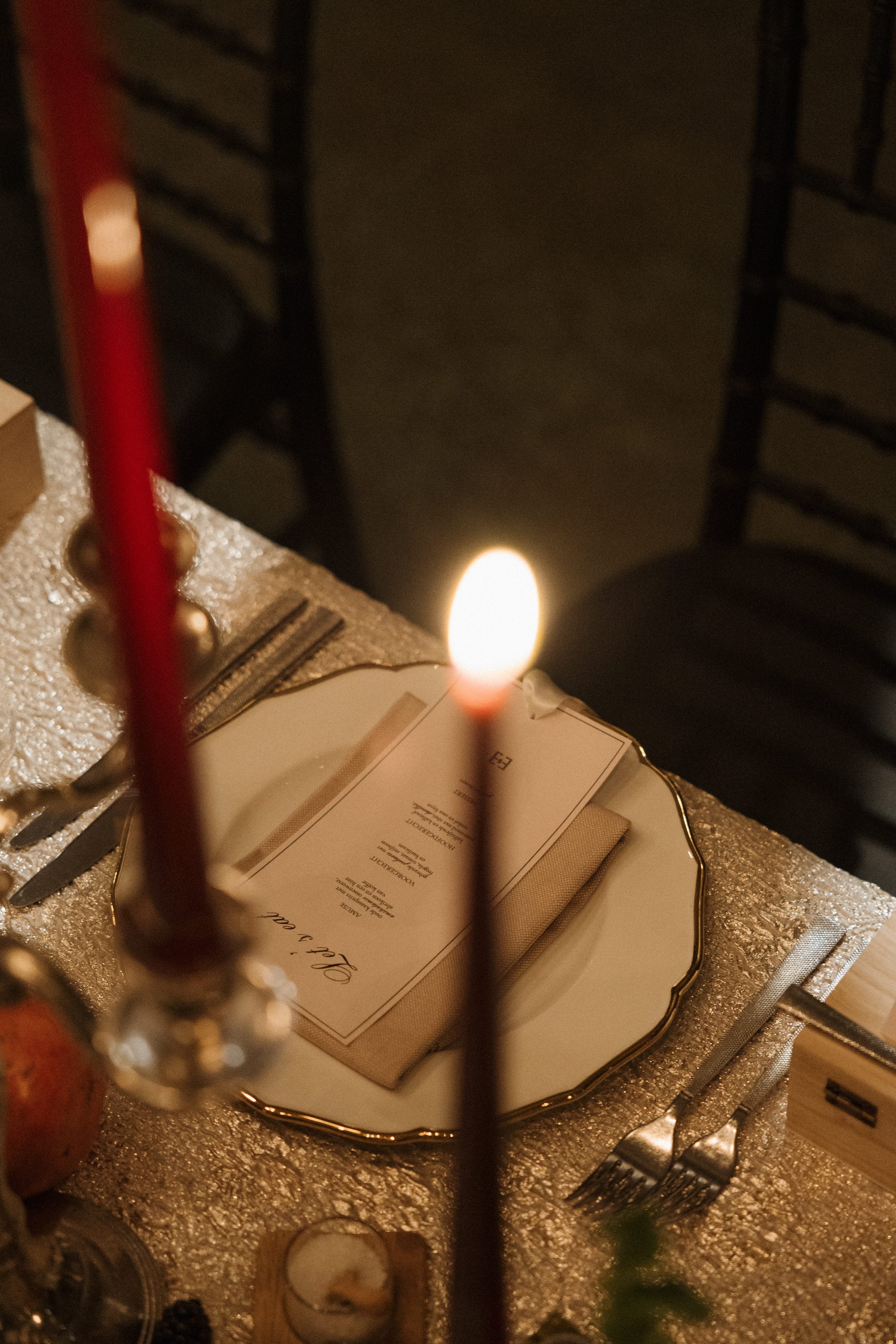 A close-up, atmospheric shot of a candlelit dinner table setting. A lit red taper candle in the foreground casts a warm glow, with another red candle in the background, a white menu card on a gold-rimmed plate, silver cutlery, and dark wooden chairs in the dimly lit room.