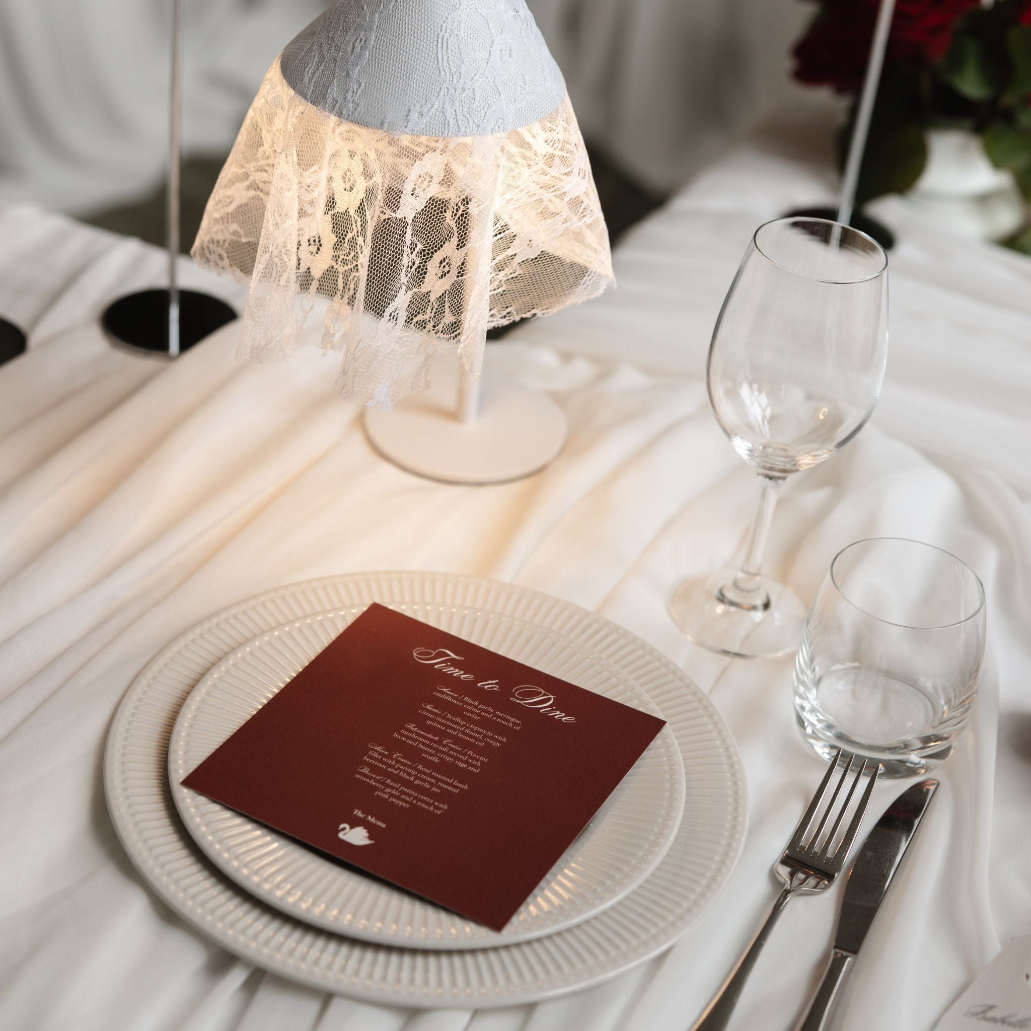 A styled table setting with white plates, a burgundy 'Time to Dine' card, clear glassware, and a decorative lamp with a lace-draped shade on a white draped tablecloth.