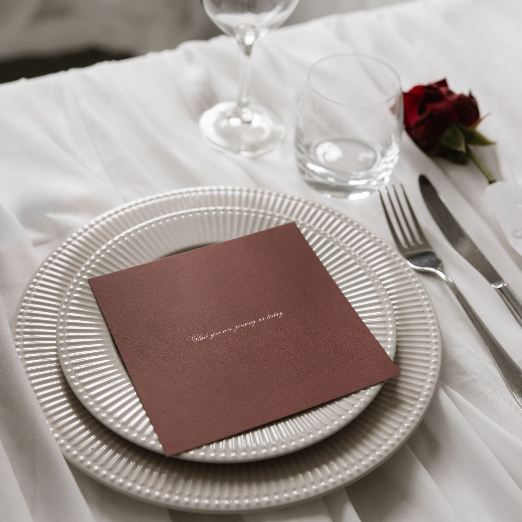 A styled place setting on a white tablecloth with two stacked white ribbed plates, a burgundy menu with white elegant text, silver flatware, a clear glass, a wine glass base and a single red rose.