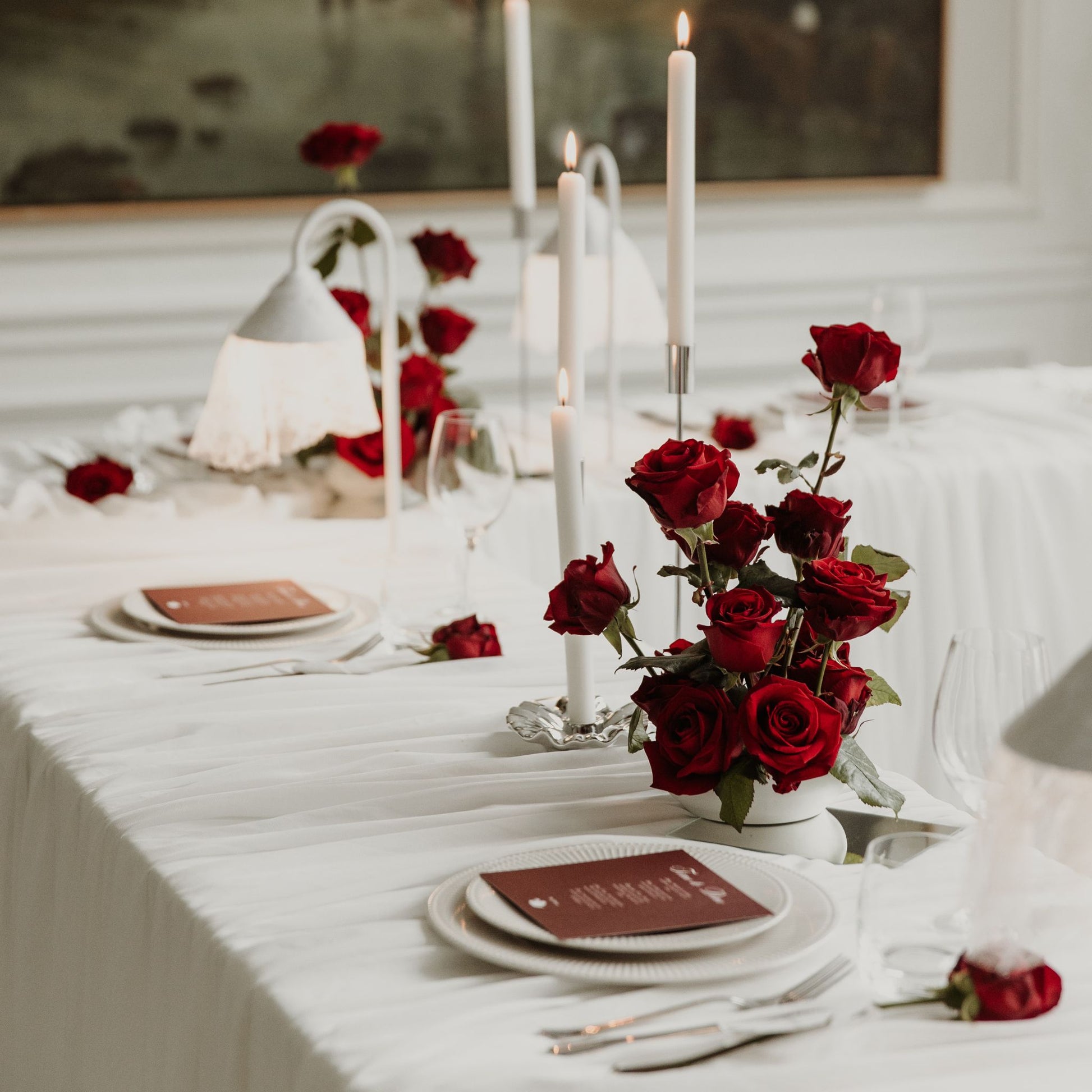 A romantic table setting draped in white linen, featuring a centerpiece of fresh red roses, tall white taper candles, and elegant place settings with bordeaux carded menus.