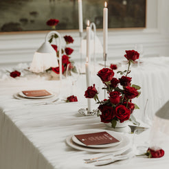 A romantic table setting draped in white linen, featuring a centerpiece of fresh red roses, tall white taper candles, and elegant place settings with bordeaux carded menus.
