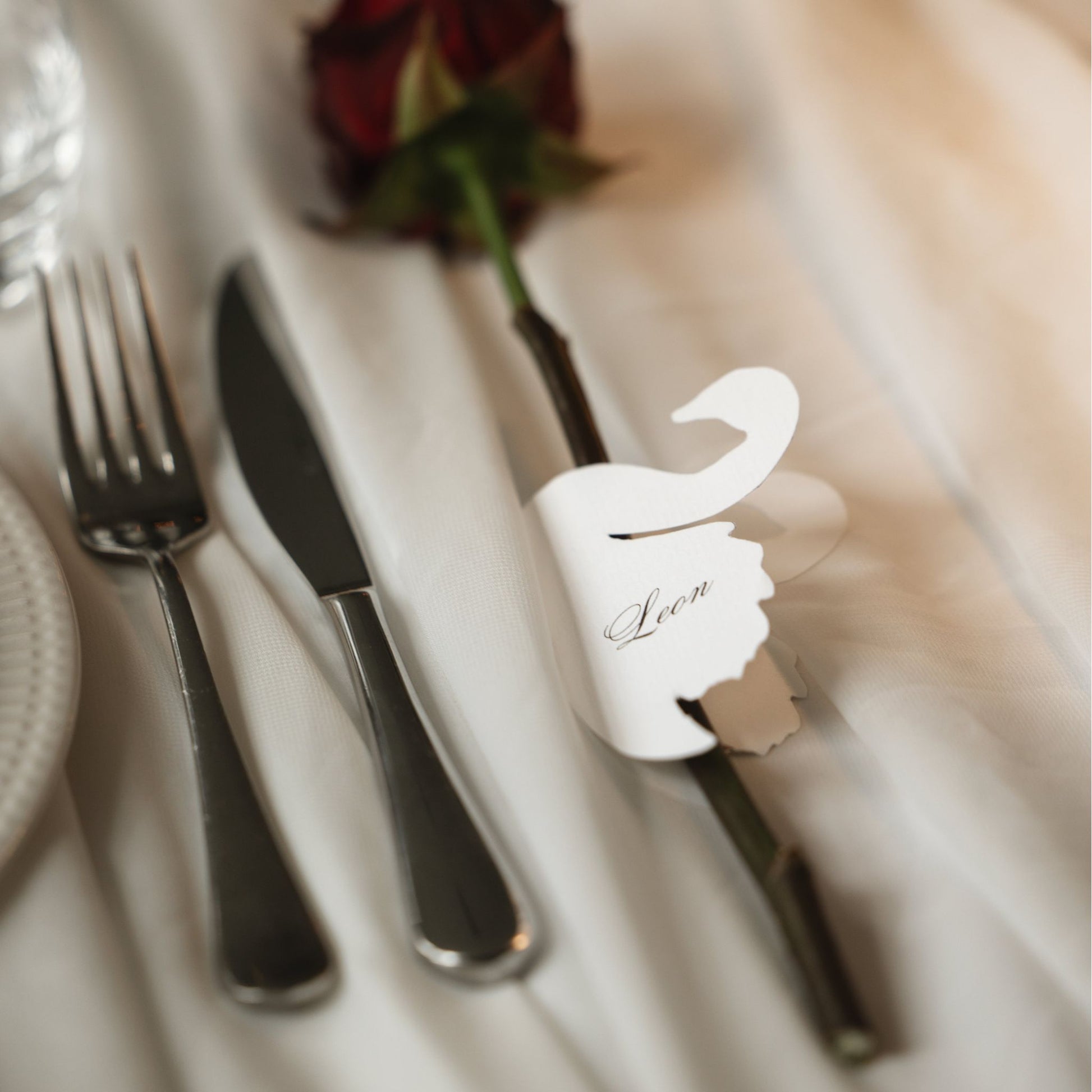 A close-up of a personalized place card holder shaped like a swan, holding a knife on a white tablecloth next to a fork and a red rose.