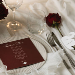 A styled wedding table setting featuring a deep burgundy place card with elegant white script reading 'Time to Dine' on a white plate, alongside silver cutlery, a white elephant name card, and a single red rose on a white tablecloth.