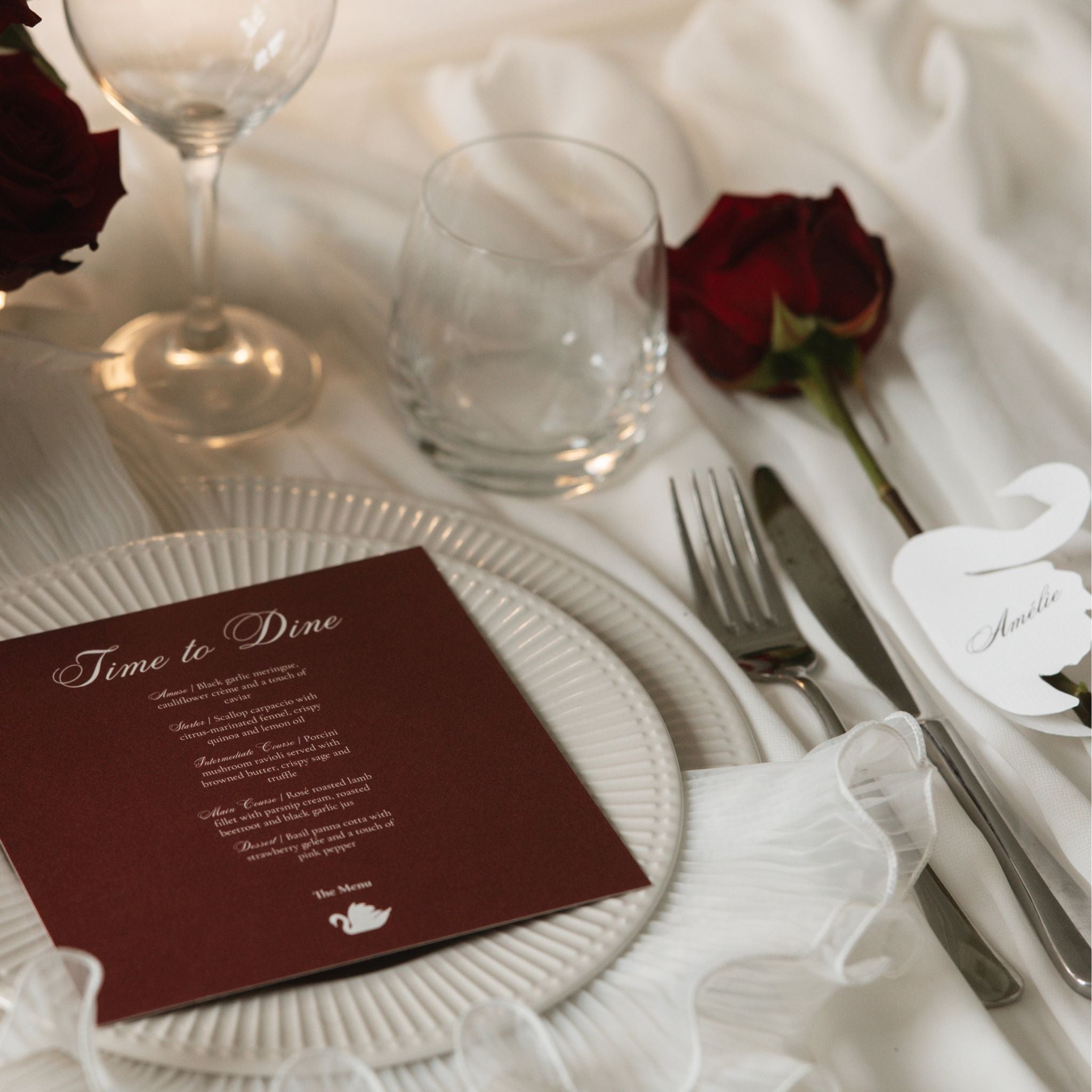 A styled wedding table setting featuring a deep burgundy place card with elegant white script reading 'Time to Dine' on a white plate, alongside silver cutlery, a white elephant name card, and a single red rose on a white tablecloth.