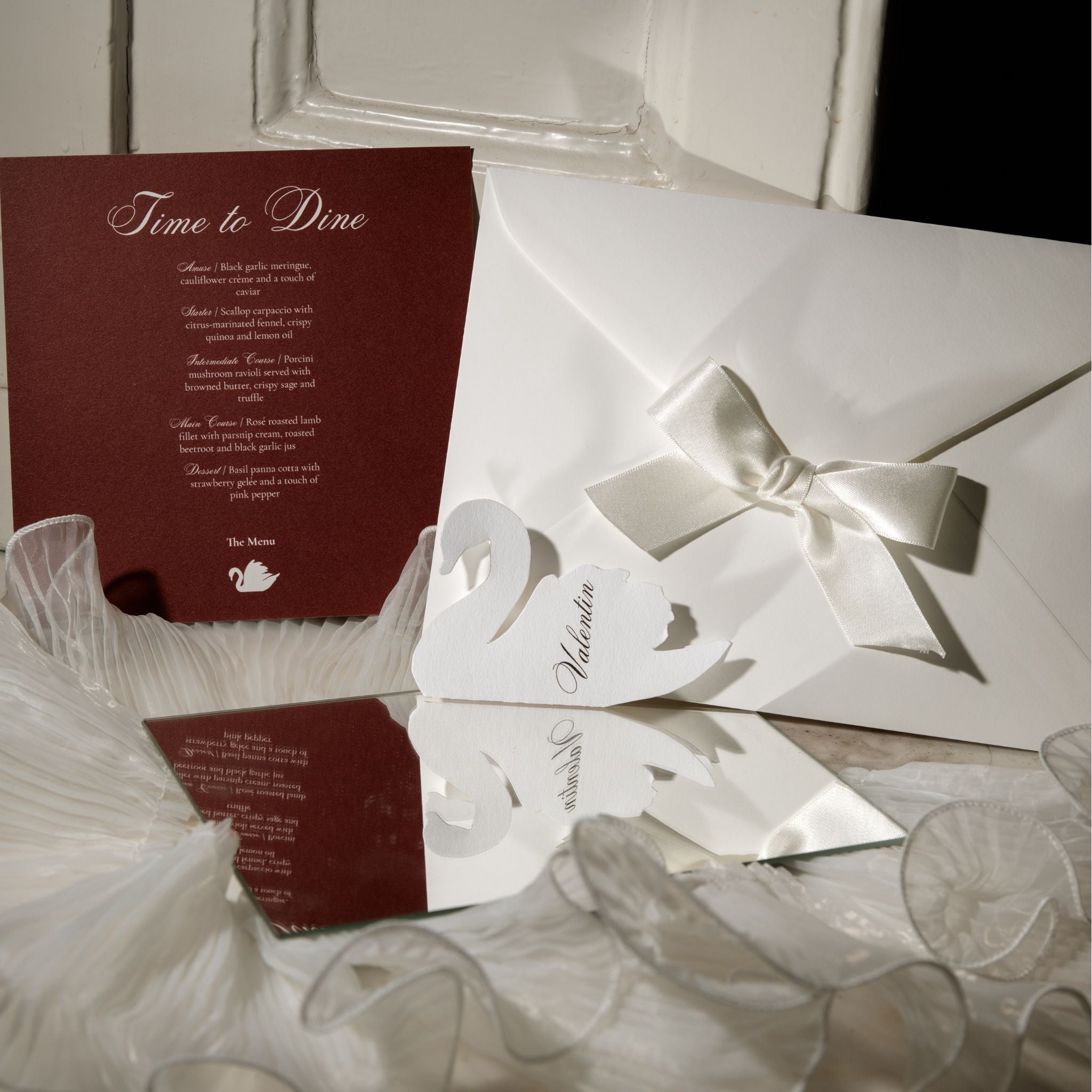 A wedding invitation suite featuring a deep burgundy card with elegant script text, a white envelope with a large white satin bow, and matching white paper cutout elements on a light surface.