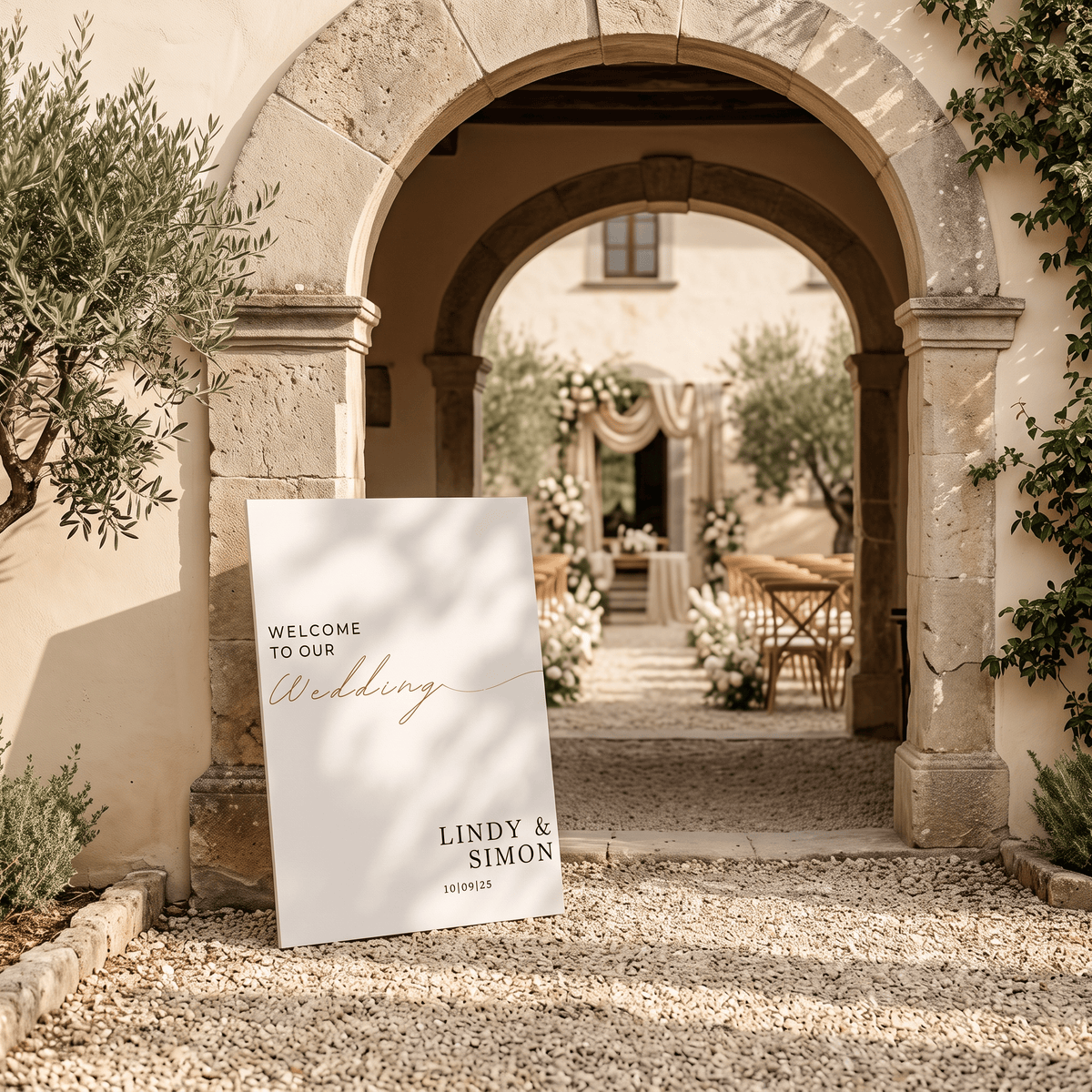 A custom wedding welcome sign for 'Lindy & Simon' stands on a gravel path outside a stone archway leading to an outdoor wedding venue. The sign features a white background with elegant black and rose gold script text.