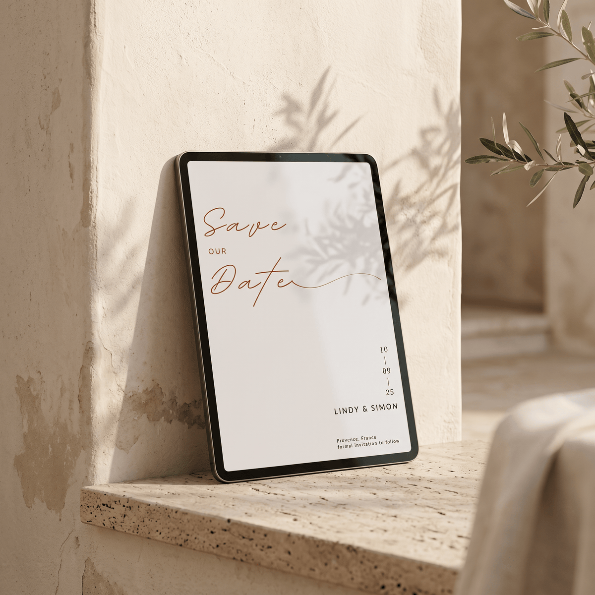 A tablet displaying a minimalist wedding save-the-date card leans against a textured beige stone wall on a matching stone ledge, with soft natural shadows and greenery in the background.