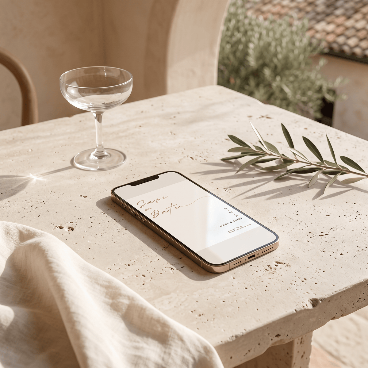 A smartphone lying on a light stone table outdoors, displaying a wedding invitation or event details screen. A clear coupe glass, a folded cream linen cloth, and an olive branch are arranged around it in a warm, sunlit setting.