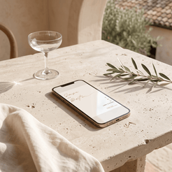 A smartphone lying on a light stone table outdoors, displaying a wedding invitation or event details screen. A clear coupe glass, a folded cream linen cloth, and an olive branch are arranged around it in a warm, sunlit setting.