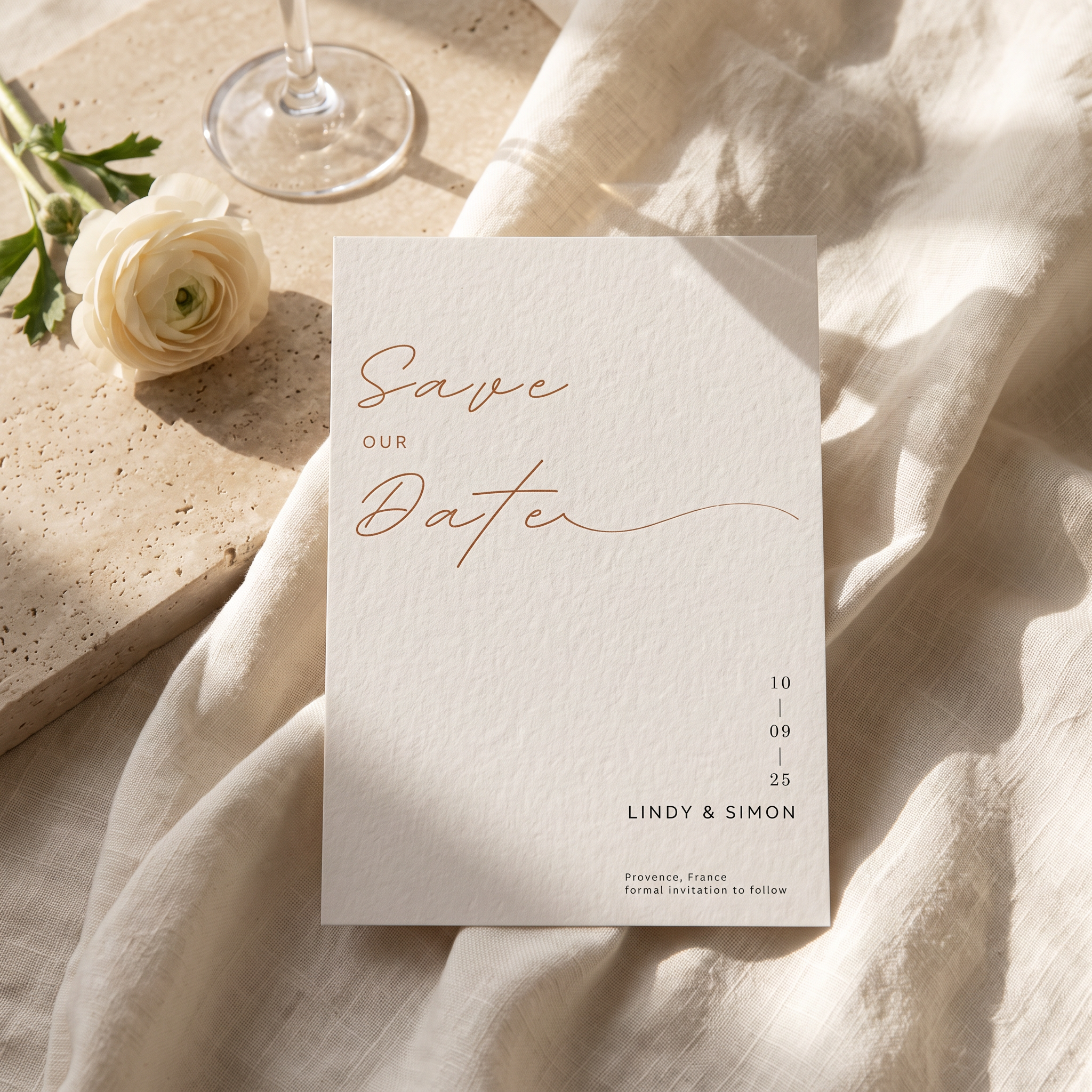 A square white wedding save-the-date card with elegant rose-gold script reading 'Save Our Date' and small black text below, styled on draped cream fabric with a stone tile, a white rose, and a wine glass in soft sunlight.