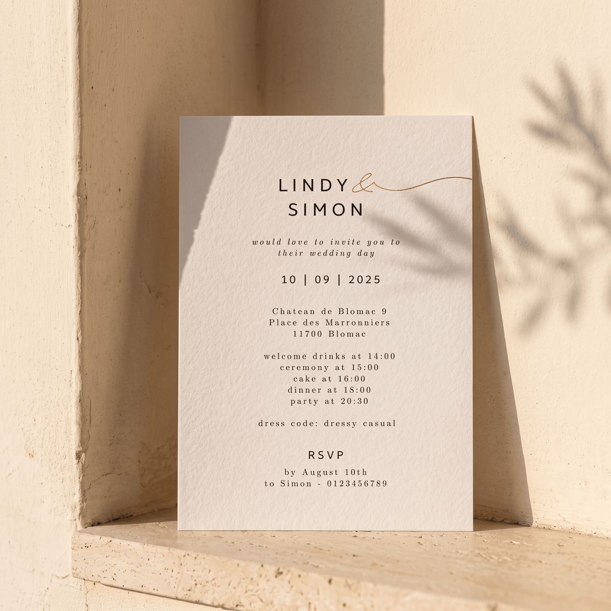 A minimalist wedding invitation card with elegant typography and a delicate gold line design, leaning against a textured beige stone wall with soft shadows cast on it.