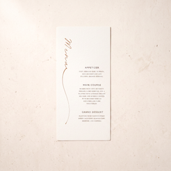 A tall, narrow white wedding menu card with elegant rose gold foil script reading 'Michelle' and black printed event details, displayed flat on a textured beige surface.