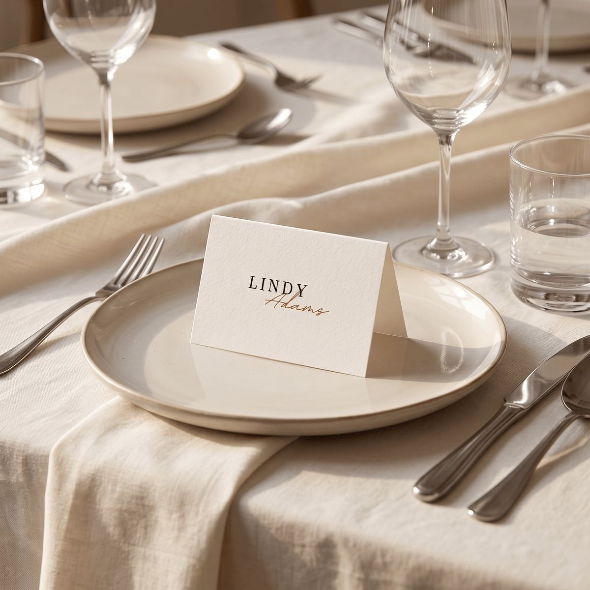 A cream-colored paper place card with the name 'LINDY Adams' printed on it, standing on a matching cream plate at a elegantly set dining table with silver cutlery, wine glasses, and water glasses.