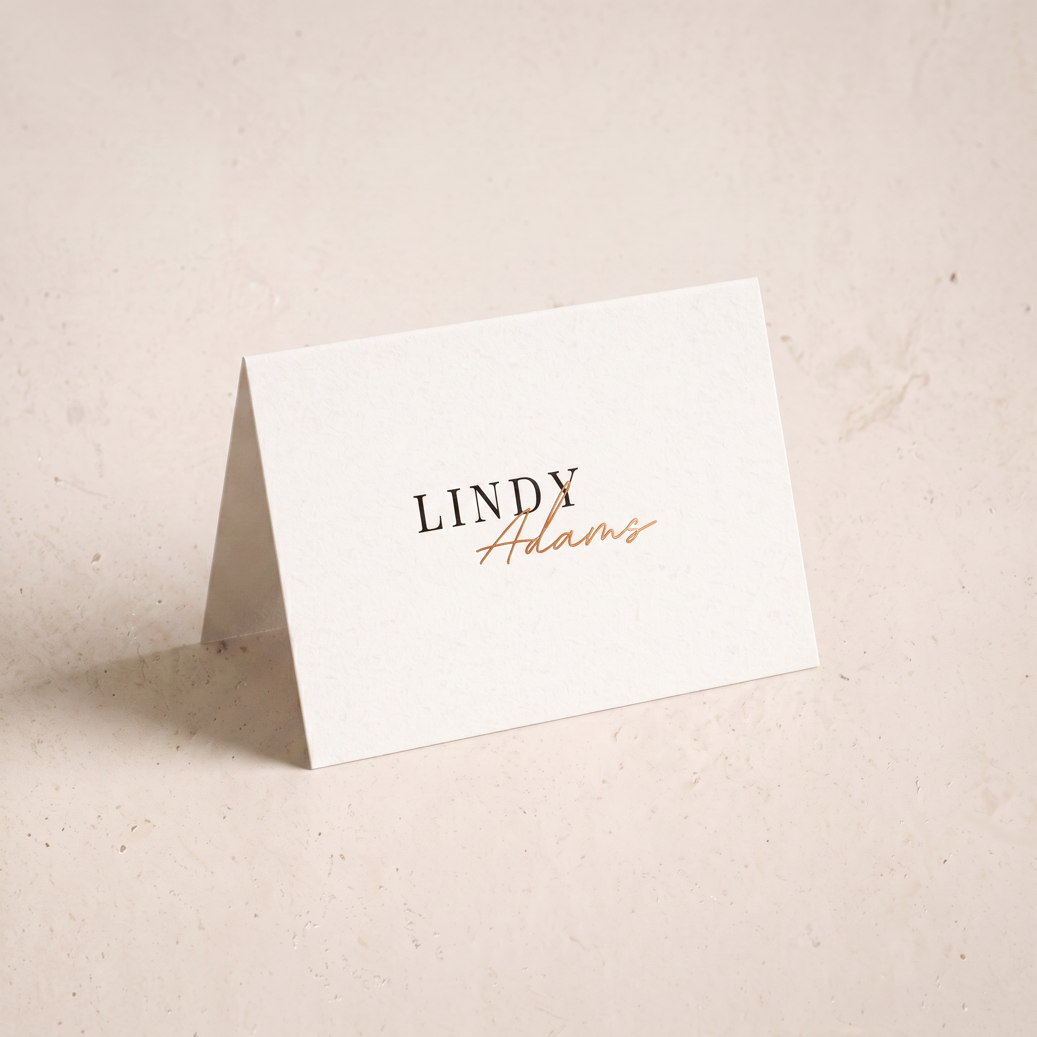 A small white standing card with the printed name 'LINDY Adams' in black and copper-gold script, photographed on a warm off-white textured surface.