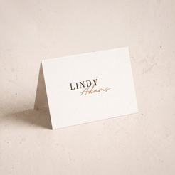 A small white standing card with the printed name 'LINDY Adams' in black and copper-gold script, photographed on a warm off-white textured surface.
