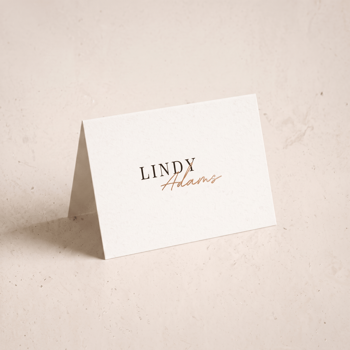 A small white standing card with the printed name 'LINDY Adams' in black and copper-gold script, photographed on a warm off-white textured surface.