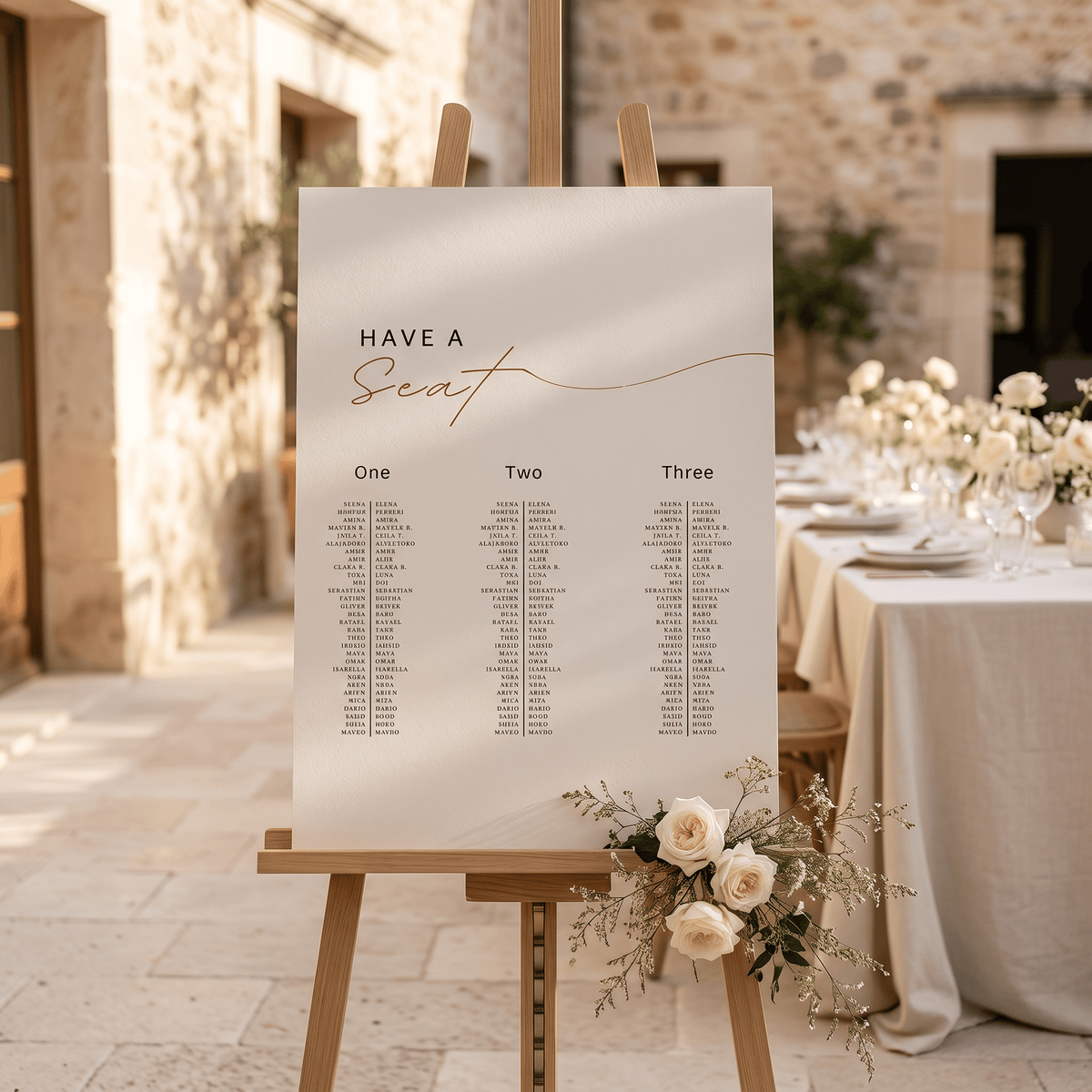 A wedding seating chart sign on a wooden easel, styled in an outdoor stone courtyard next to a decorated event table with white flowers and table linens.
