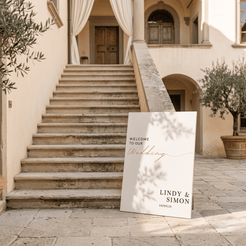 A custom wedding welcome sign leaning against a stone staircase in a sunlit courtyard. The sign is off-white with elegant black serif text reading 'WELCOME TO OUR Wedding' and names 'LINDY & SIMON'.