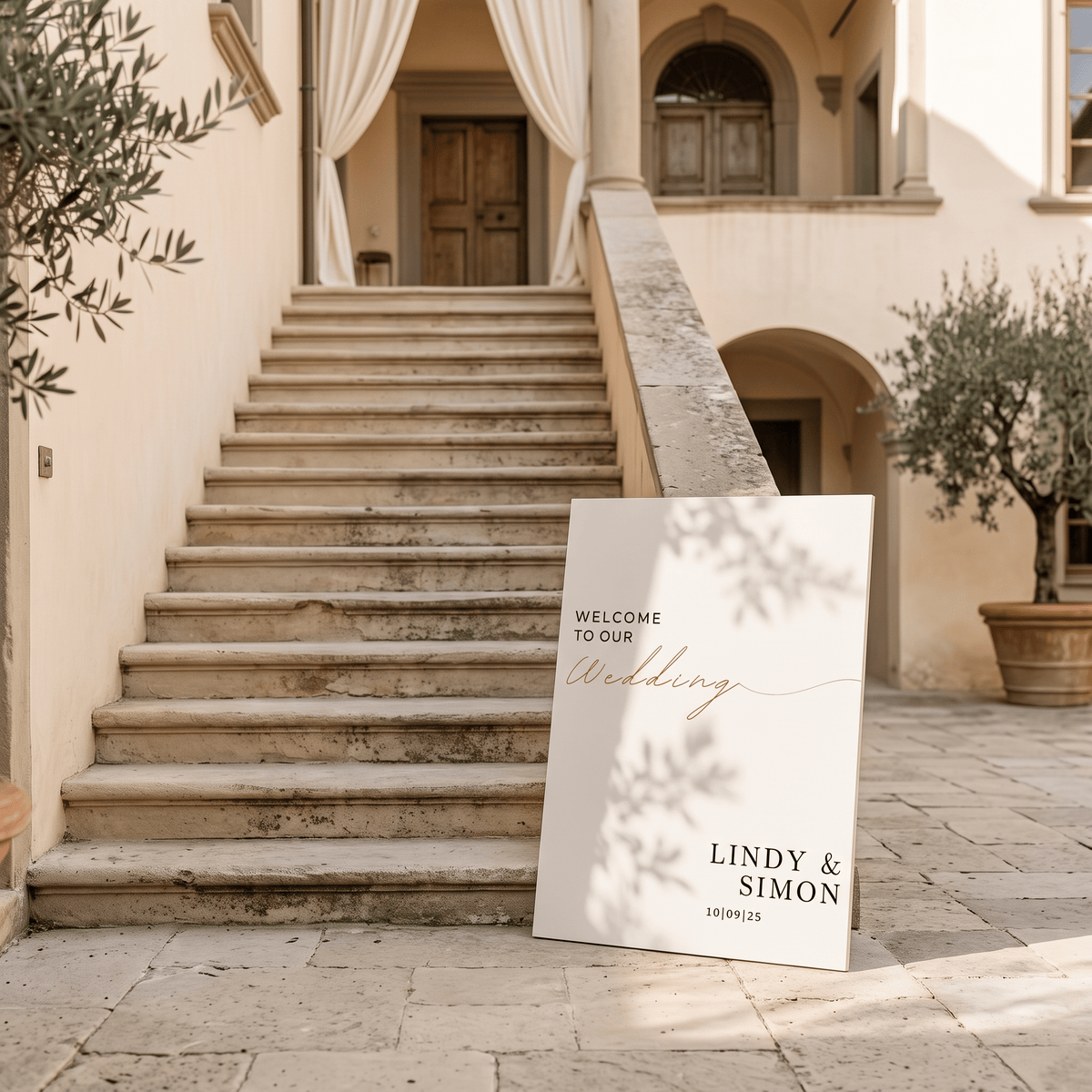 A custom wedding welcome sign leaning against a stone staircase in a sunlit courtyard. The sign is off-white with elegant black serif text reading 'WELCOME TO OUR Wedding' and names 'LINDY & SIMON'.