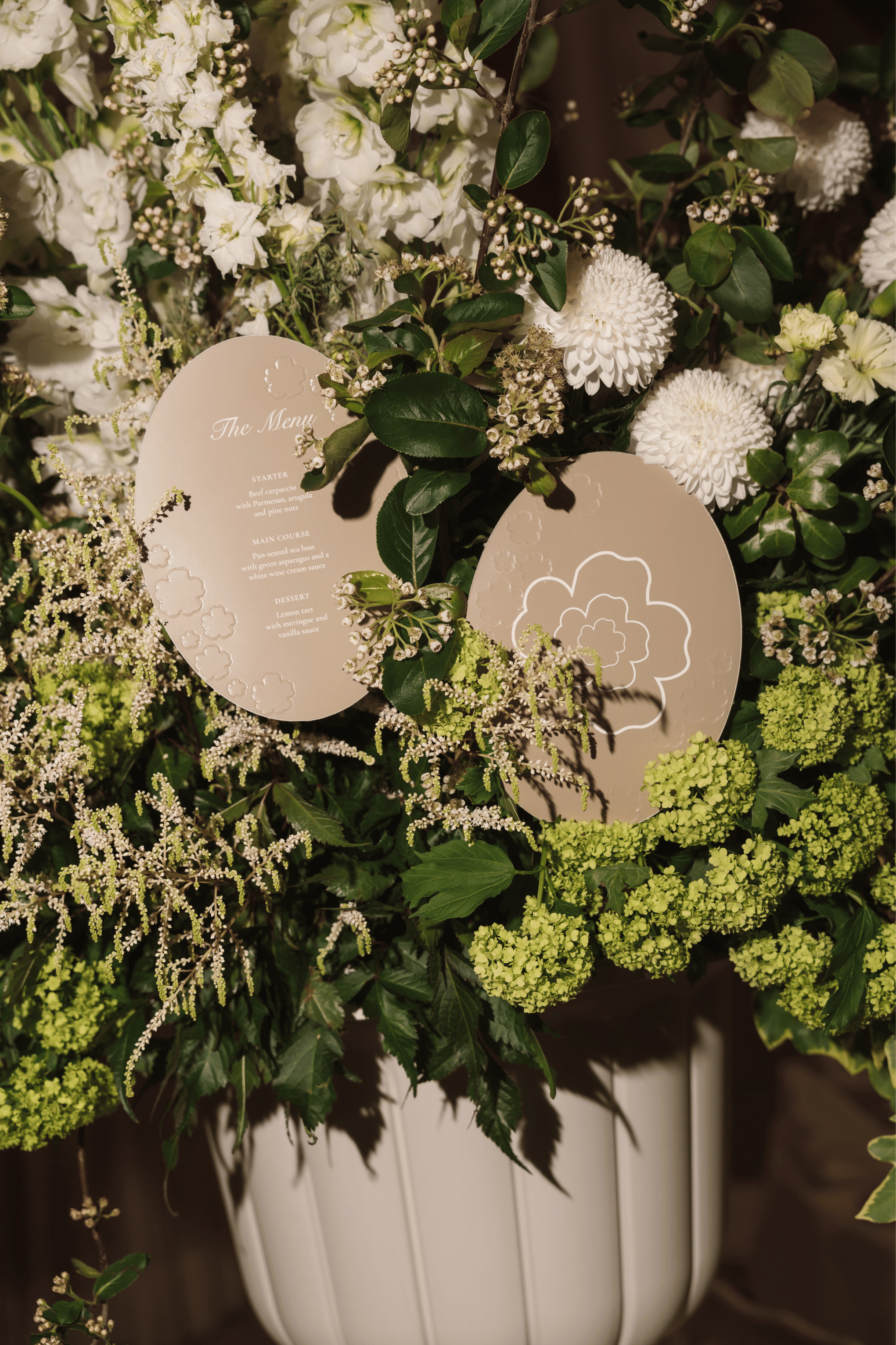 Two round beige paper place cards with elegant printed designs, nestled inside a large white floral arrangement with white and green flowers.