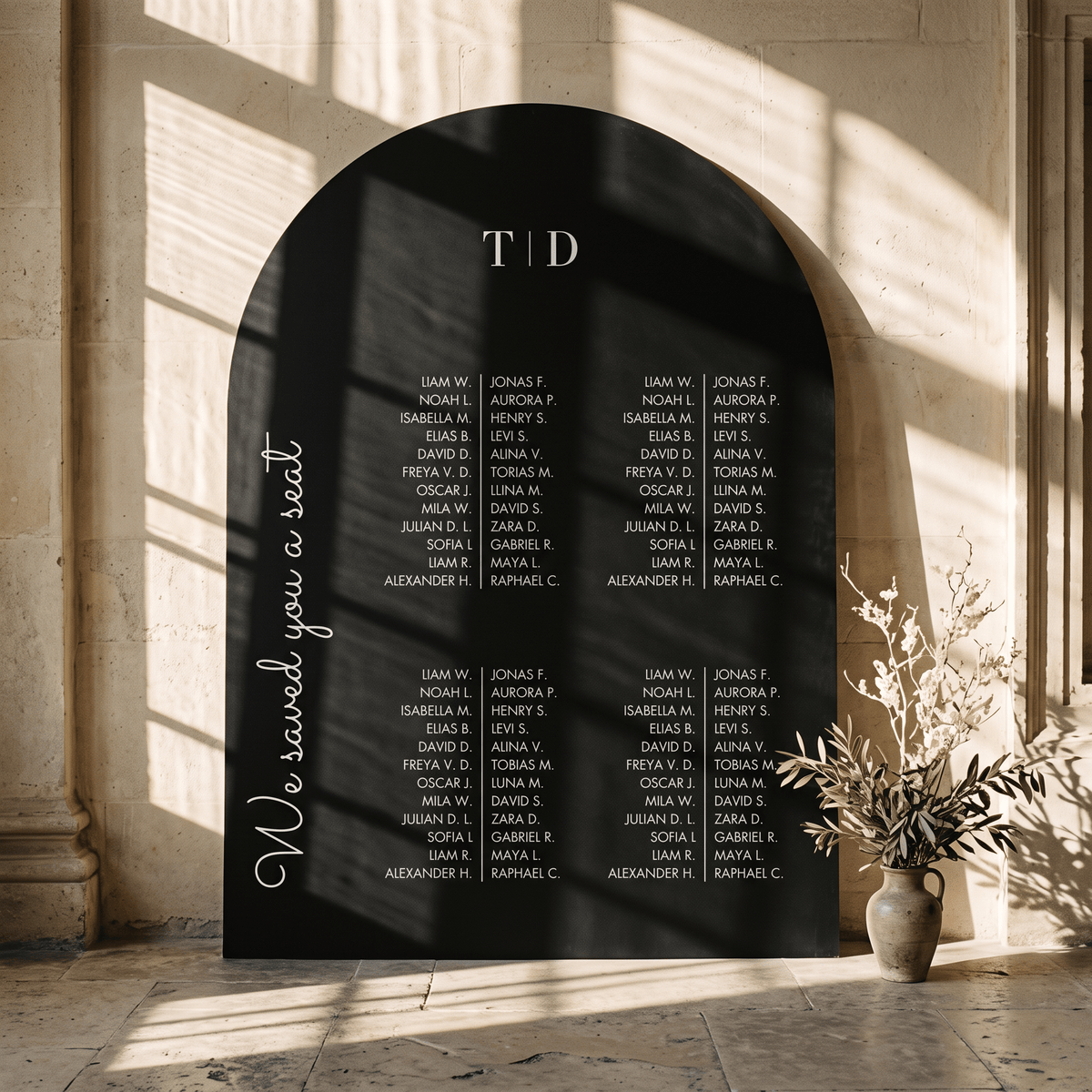 An arched black wedding seating sign with white printed names and the words 'We saved you a seat' and 'TID', displayed in a sunlit stone interior with a vase of branches nearby.