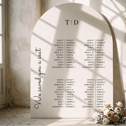 A large arched white wedding seating sign displayed in a sunlit stone interior. The sign reads 'We saved you a seat' and lists names under the heading 'TID'.