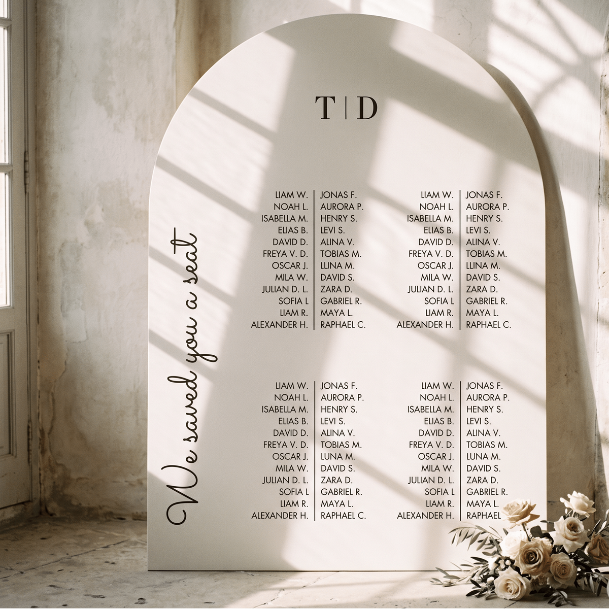 A large arched white wedding seating sign displayed in a sunlit stone interior. The sign reads 'We saved you a seat' and lists names under the heading 'TID'.