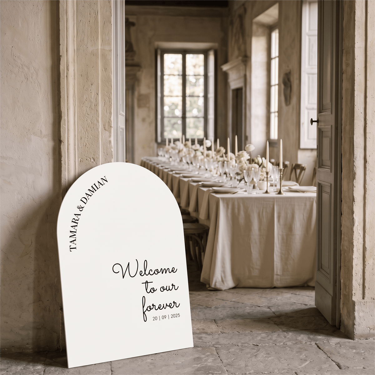 A white arched welcome sign with black script text reading 'Welcome to our forever' and names/date printed on it, leaning against a stone wall in a real indoor event space with a long table set in the background.